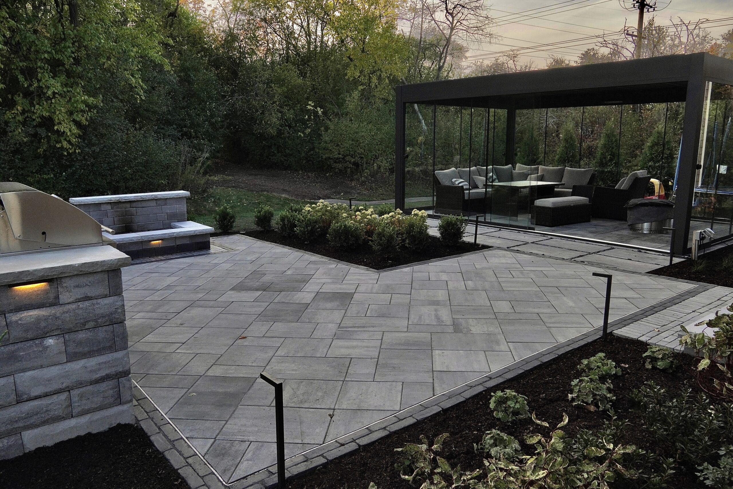 Modern Paver Patio with Glass Pavilion Modern paver patio with integrated lighting, outdoor kitchen, and glass-enclosed pavilion.